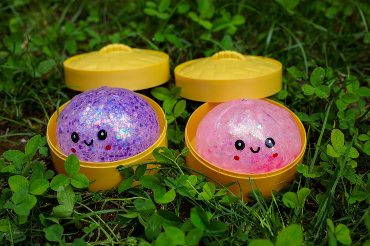 Two glitter squishy dumpling toys in purple and pink sitting in open yellow steamer containers